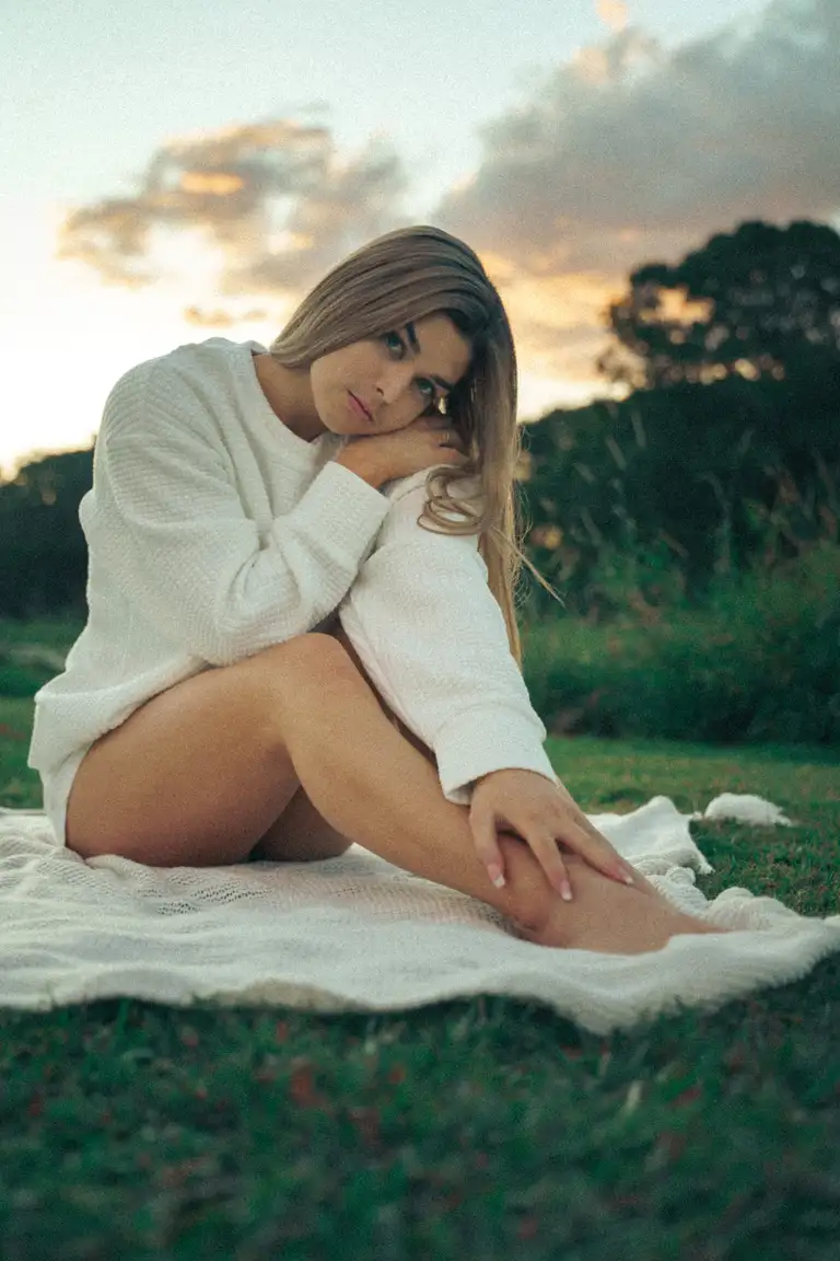 Soft lifestyle fashion portrait of a model sitting on a blanket at sunset in Australia, wearing a cozy white knit jumper with warm, cinematic tones and relaxed moo