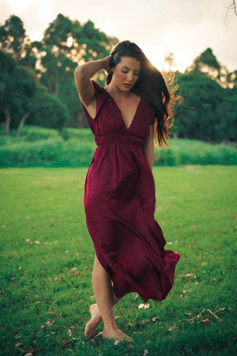 Editorial fashion image of a model in a flowing deep-red maxi dress walking barefoot in a grassy park in South East Queensland, photographed with soft tones and graceful movement.