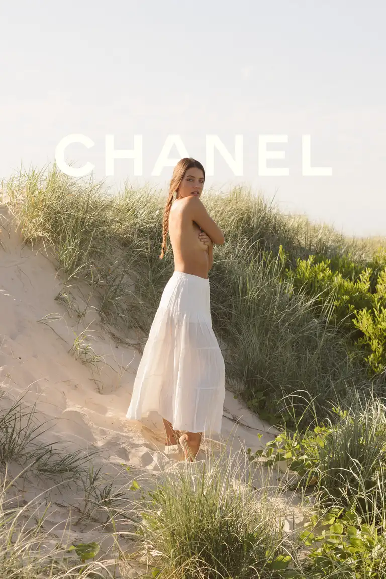 Commercial fashion photograph of a model wearing a white flowing maxi skirt, posed topless in sunlit coastal sand dunes with a natural boho aesthetic and soft film-look styling, showcasing the garment’s movement and texture.