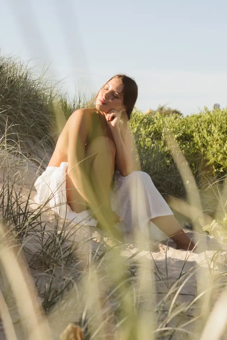 Model seated in coastal sand dunes wearing a white boho-style skirt, captured in warm golden hour light with a relaxed, dreamy film-look aesthetic to highlight the skirt’s soft fabric and lifestyle appeal.