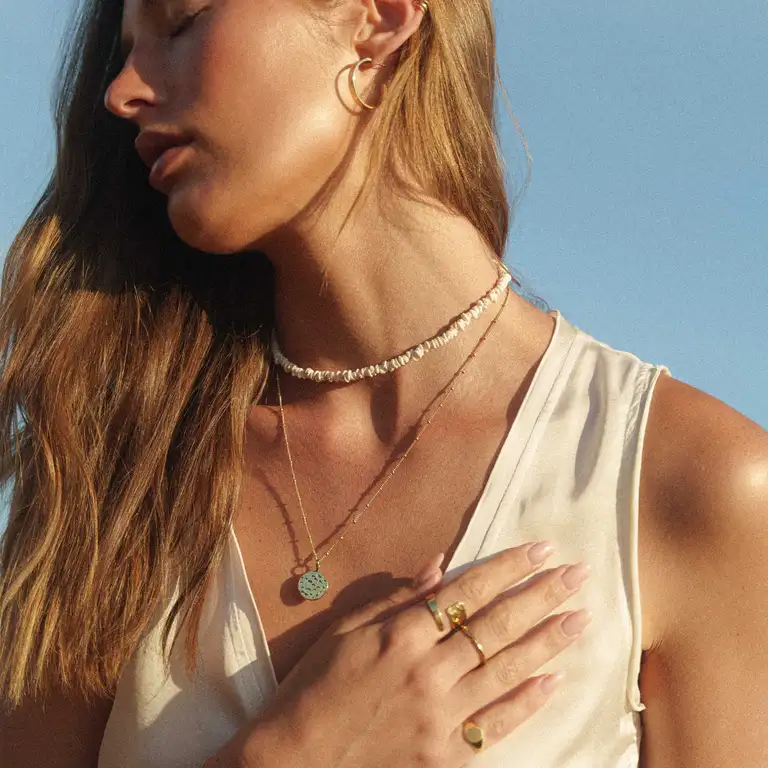 Golden hour close-up portrait of a model wearing layered gold necklaces and rings from Aquila Jewellery, styled in a boho, film-look aesthetic against a clear sky.