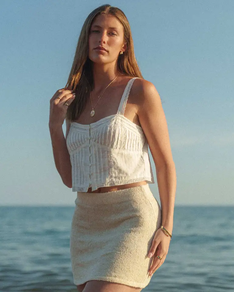 Mid shot of model wearing a white top and skirt at the beach in golden hour light, featuring boho-inspired jewellery from a jewellery brand.