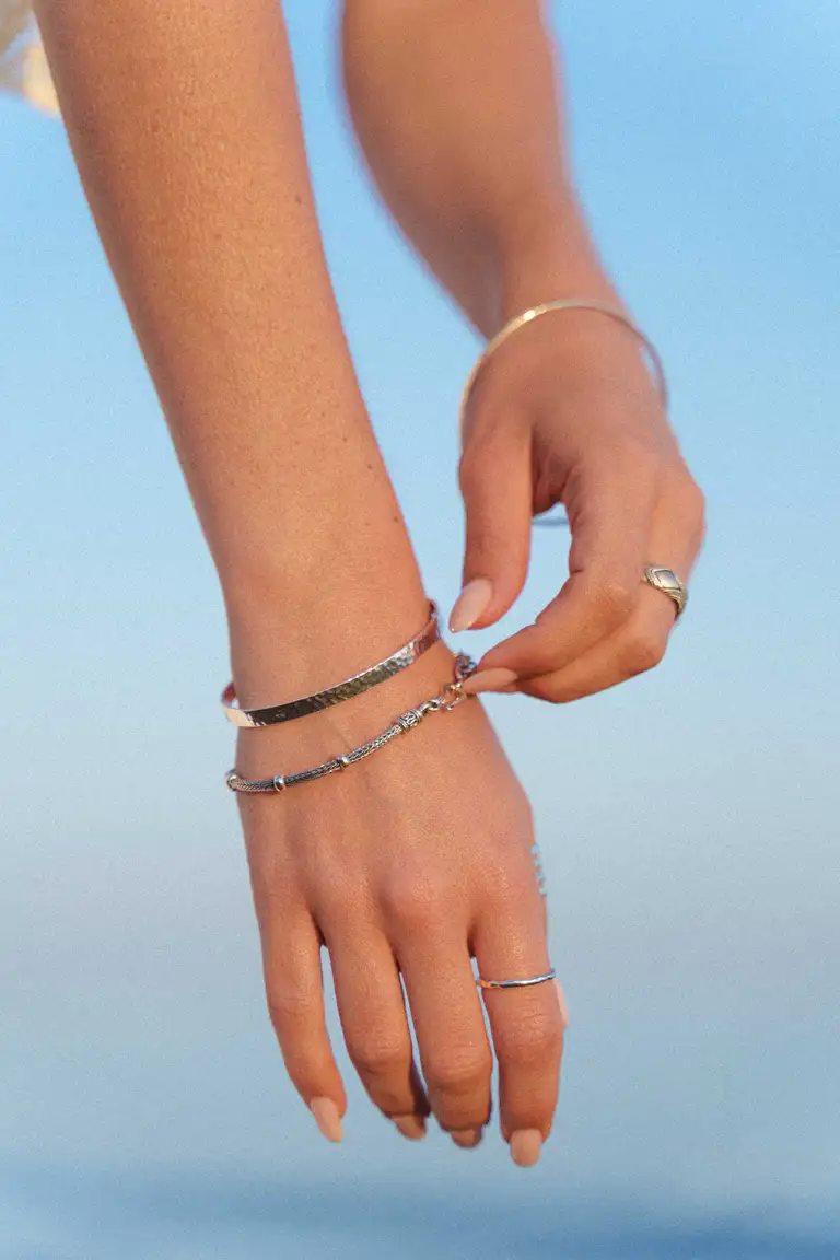 Close-up of hands wearing gold rings and bracelets from a jewellery brand, styled in a relaxed boho look against a coastal golden hour backdrop.