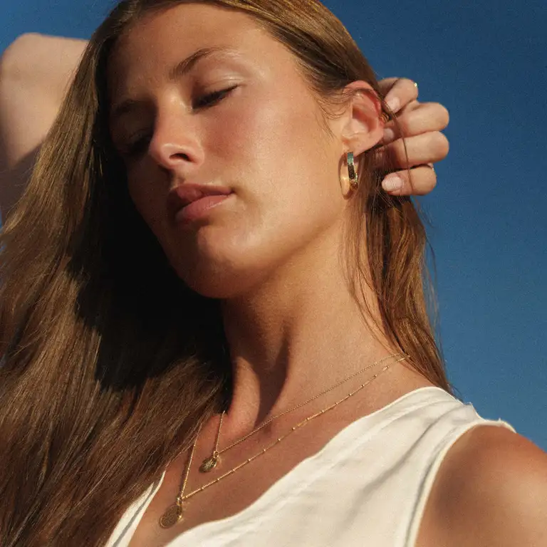 Boho-style portrait of a model in golden hour sun, eyes closed, wearing minimalist gold jewellery from Aquila Jewellery with a dreamy film-look aesthetic.