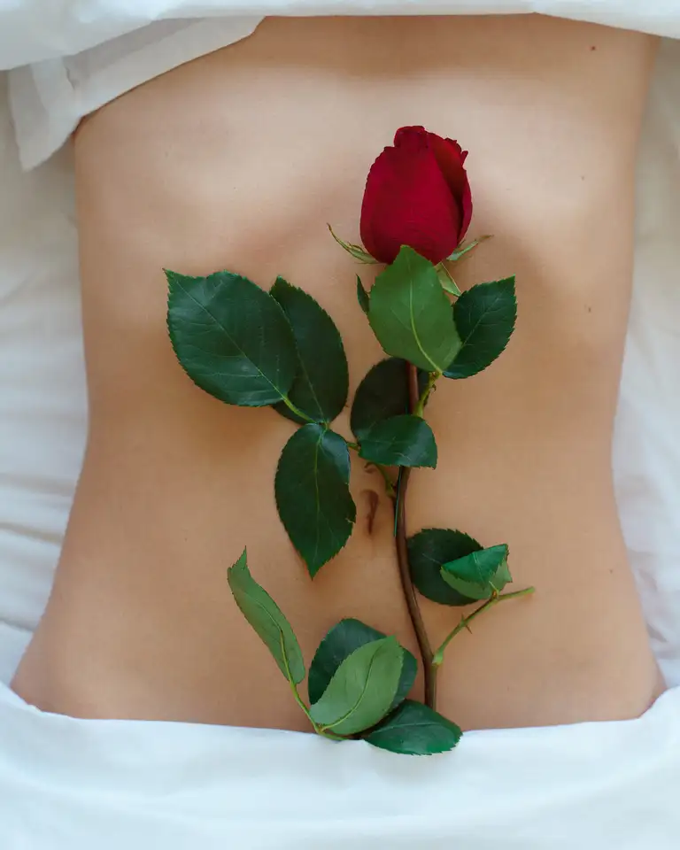 Artistic photo of a rose laying on the torso of a woman who is on a bed with white sheets, photographed in soft daylight on the Gold Coast in the home studio of Bora Mustafa.