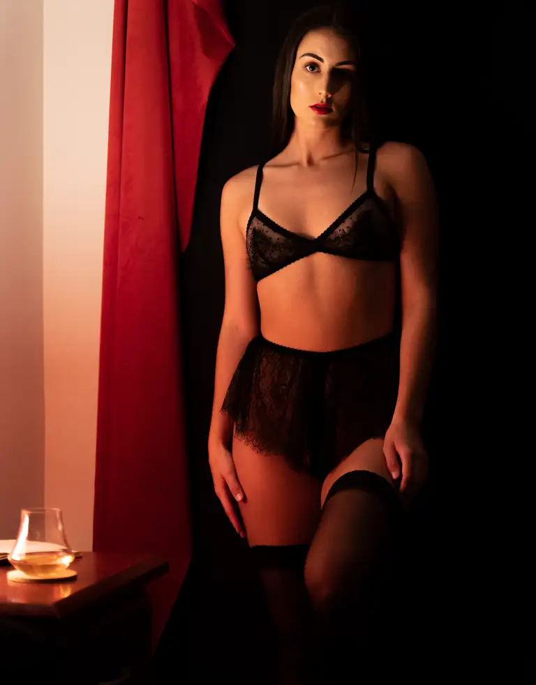 Model standing beside red curtains wearing a black lace lingerie set from Lazy Girl Lingerie, photographed in moody candlelight on the Gold Coast for a commercial lingerie shoot.ye